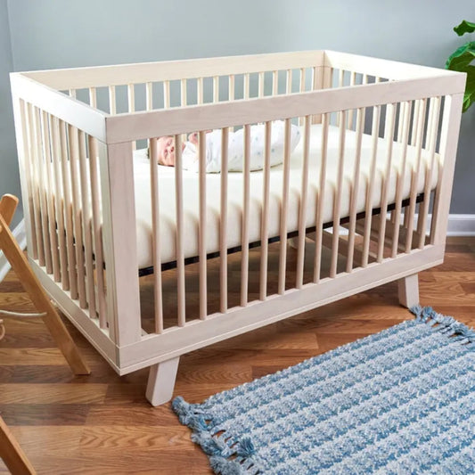 White crib with a baby inside on a wooden floor with a blue rug