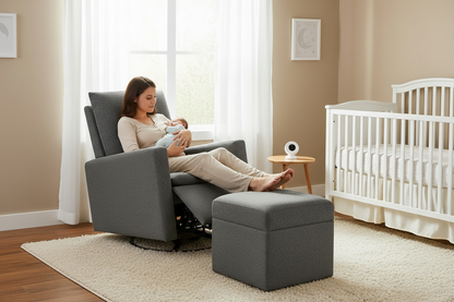 A woman in a beige outfit sits in a gray upholstered recliner chair, holding a baby wearing a light blue outfit. A matching gray storage ottoman is in front of the chair, and the room includes a white crib, a small wooden side table with a white device, beige walls, and a cream rug.
