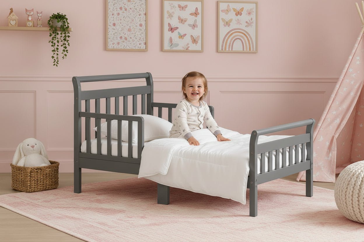 A gray toddler bed with safety rails and a white mattress, styled in a nursery setting with a teddy bear basket, framed wall art, and a teepee tent.