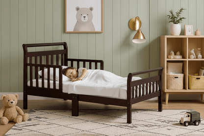 A toddler bed with dark brown wooden rails and a safety guard, shown with a child sleeping underneath a white blanket in a styled children's bedroom.