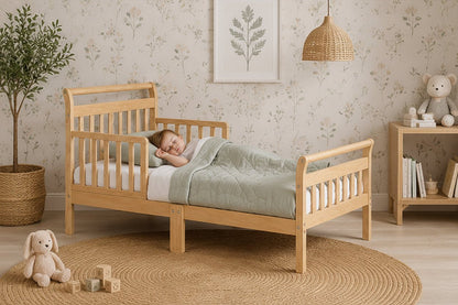 A light natural wood toddler bed with safety rails, styled in a cozy children's bedroom with floral wallpaper, a woven rug, a teddy bear, and a small wooden shelf.