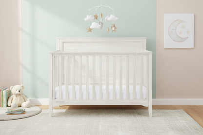 White crib in a nursery with a teddy bear and decorative items on a light green wall.