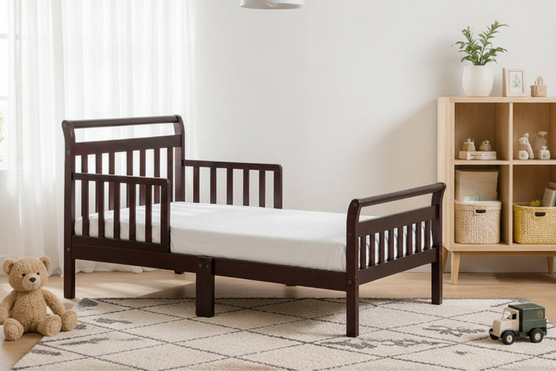 A dark brown wooden toddler bed with guard rails and a side panel, shown in a styled children's bedroom with a green paneled wall, wooden shelf, teddy bear, and wall lamp.