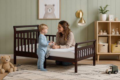 A dark brown wooden toddler bed with guard rails and a side panel, shown in a styled children's bedroom with a green paneled wall, wooden shelf, teddy bear, and wall lamp.