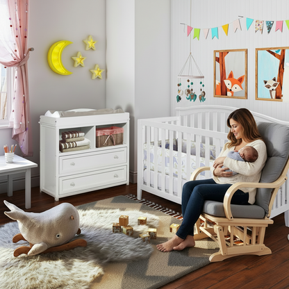 A woman in a rocking chair holds a baby in a nursery setting. The room features a white changing table, a white crib, a gray and beige rug, a plush whale toy, and decorative wall accents including a yellow crescent moon and star lights.