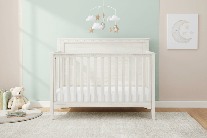 White crib in a nursery with a teddy bear and decorative items on a light green wall.