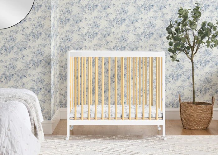 White crib with wooden slats in a room with floral wallpaper and a plant.