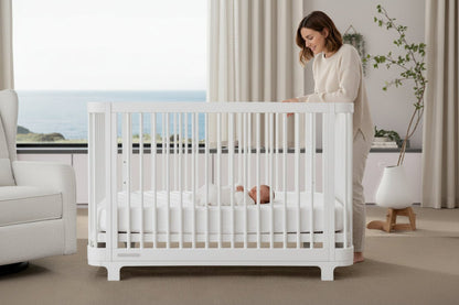 White crib with a blue mattress in a room with large windows and curtains.