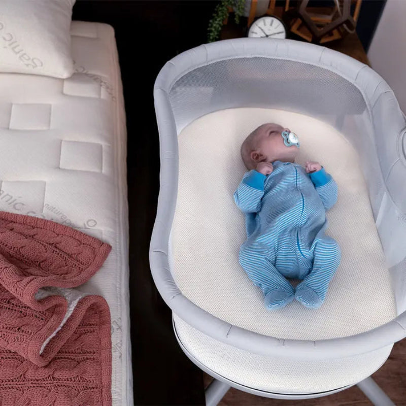 Baby in a blue outfit lying in a white bassinet next to pink towels.