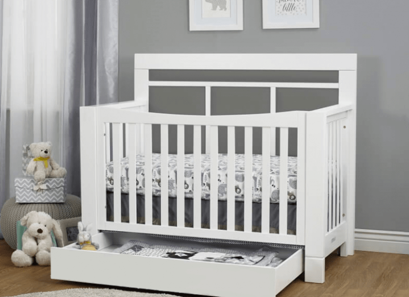 White crib with a gray mattress in a room with gray walls and white curtains.