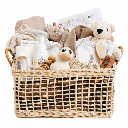 A wicker baby gift basket filled with soft baby items including a teddy bear, plush duck, onesie, bottles, and folded blankets, photographed on a clean white background.