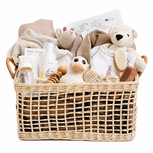 A wicker baby gift basket filled with soft baby items including a teddy bear, plush duck, onesie, bottles, and folded blankets, photographed on a clean white background.